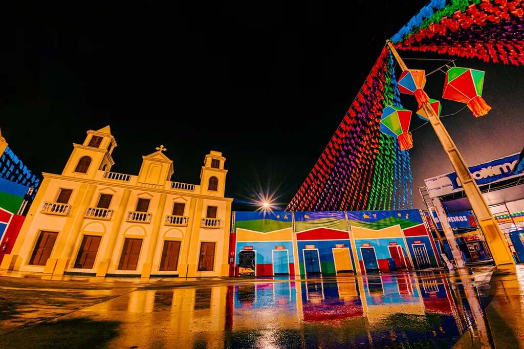 Parque do Povo com um edifício clássico e construções coloridas ao redor, além de bandeirinhas de festa junina e balões de São João pendurados no poste.